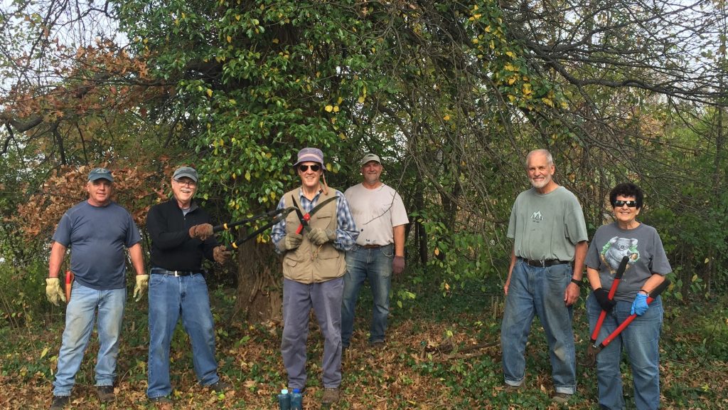 Six adults stand in a line outdoors, holding pruning tools in a leafy backyard setting.